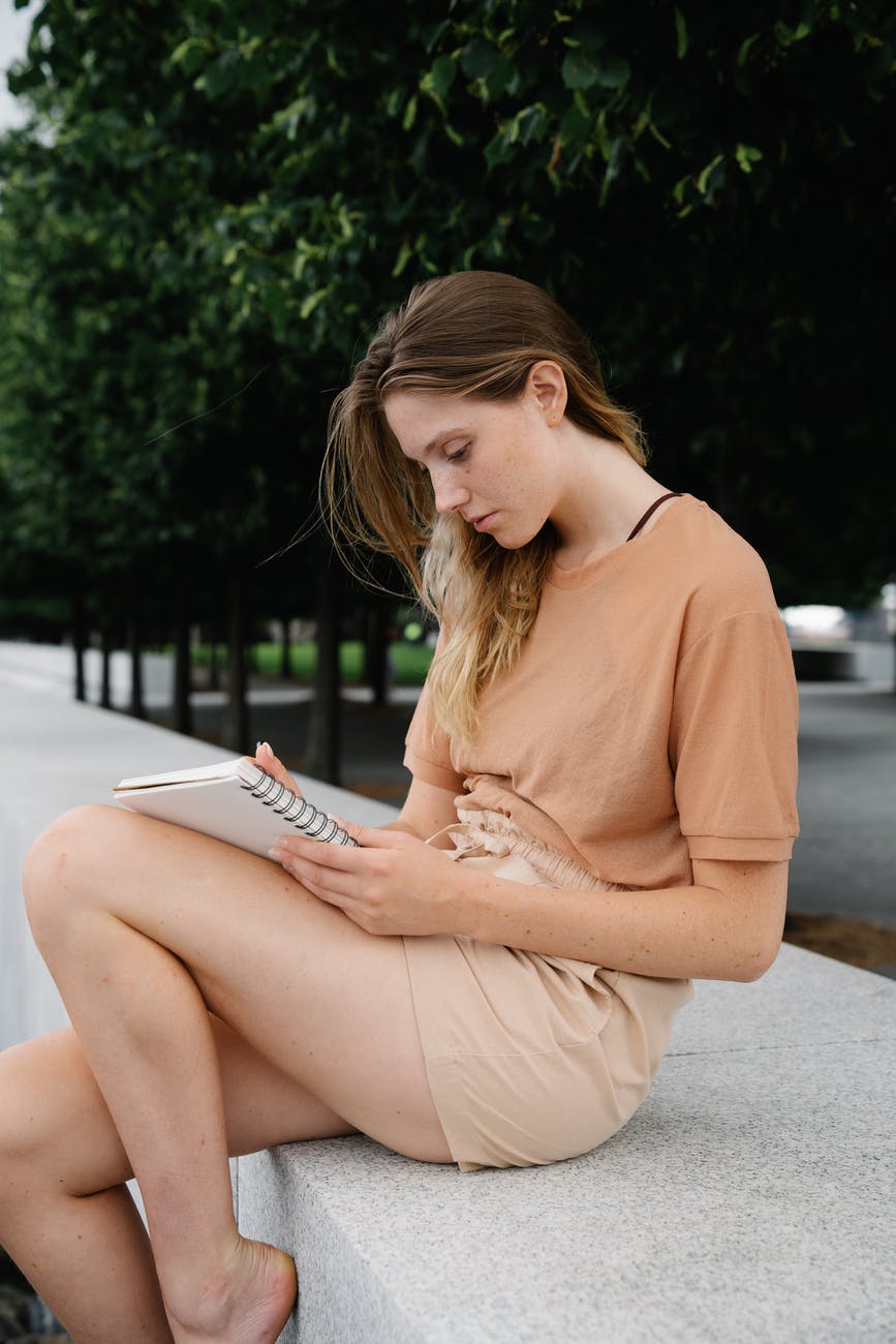 woman in brown shirt sitting on a concrete surface while writing on a notebook. love yourself